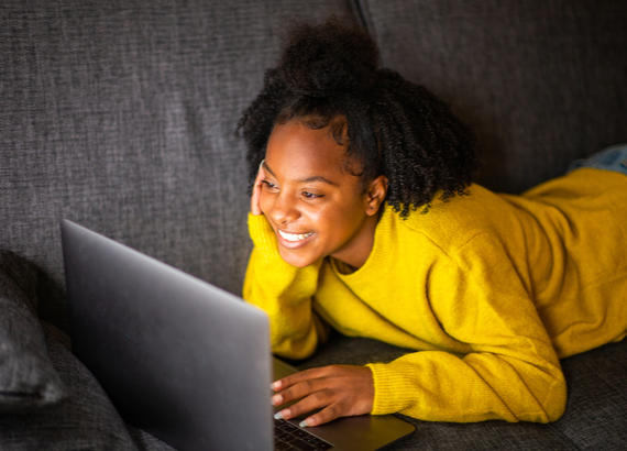 Teen girl laying on couch, resting head on hand, smiling at computer